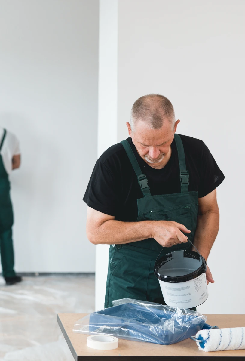 Maler bei Innen-Malerarbeiten gießt graue Farbe aus Eimer auf Malervlies neben Farbrolle und Kreppband