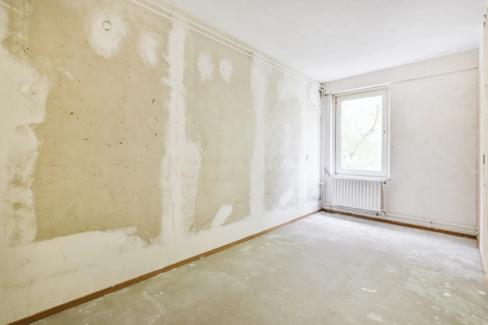 Unfertiger Raum mit gespachtelten Wänden, Estrichboden, Fensterbank und Heizkörper im Zuge von Spachtelarbeiten