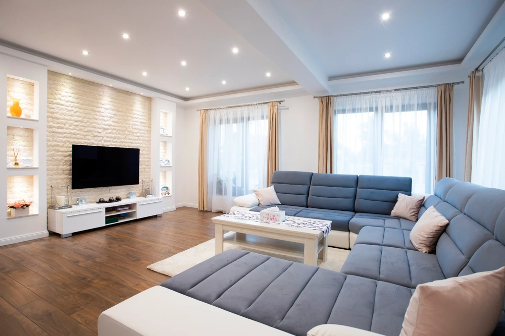 Helles Wohnzimmer mit abgehängter Decke, eingelassener Beleuchtung, graublauer Polster-Sitzlandschaft und Holzparkett