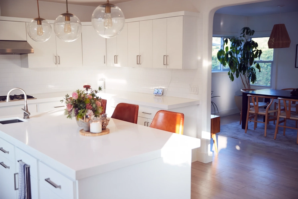 Moderne weiße Kücheninsel mit hochwertiger Arbeitsplatte, Lederbarhockern, Hängelampen und angrenzendem Essbereich – Küchenrenovierung